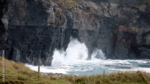 White waves clashing against a dark rocky cliff  with some grass in the foreground.