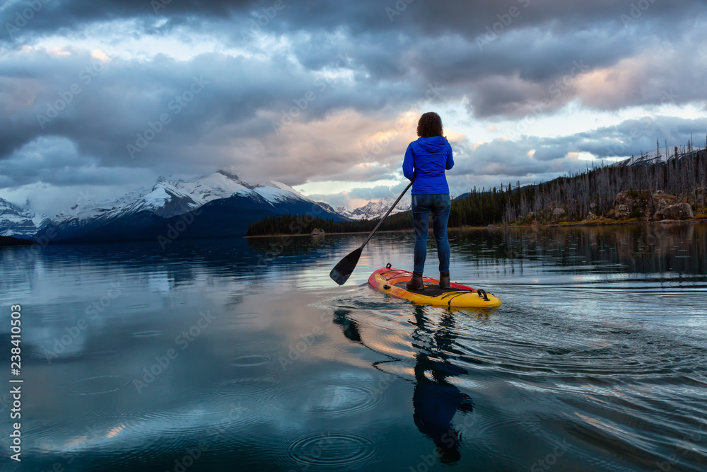Girl Paddle Boarding in a peaceful and calm glacier lake during a vibrant cloudy sunset. Taken in Maligne Lake, Jasper National Park, Alberta, Canada.