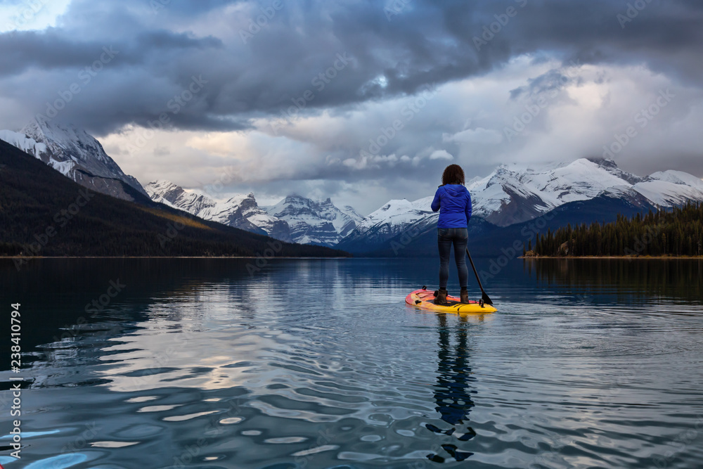Naklejka premium Girl Paddle Boarding in a peaceful and calm glacier lake during a vibrant cloudy sunset. Taken in Maligne Lake, Jasper National Park, Alberta, Canada.