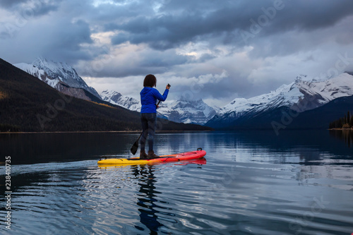Girl Paddle Boarding in a peaceful and calm glacier lake during a vibrant cloudy sunset. Taken in Maligne Lake  Jasper National Park  Alberta  Canada.