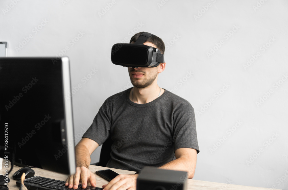 Young man wearing virtual reality goggles headset, vr box and sitting in the office against computer monitor. Connection, technology, new generation, progress concept.
