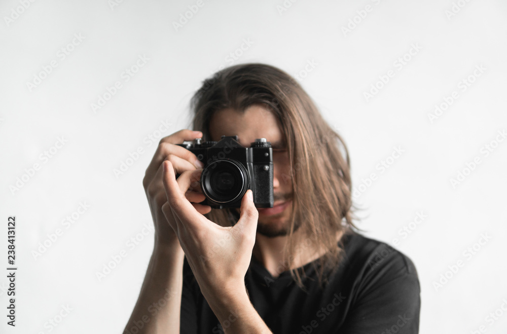 Handsome young bearded man with a long hair and in a black shirt holding vintage old-fashioned film camera on a white background and looking in camera viewfinder.