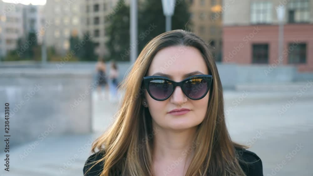 Portrait of young businesswoman in sunglasses walking in city street. Attractive business woman looking at the camera. Face of confident girl commuting to work. Close up Slow motion
