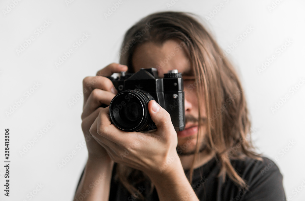 Handsome young bearded man with a long hair and in a black shirt holding vintage old-fashioned film camera on a white background and looking in camera viewfinder. Handsome young bearded man with a long hair and in a black shirt holding vintage old-fashioned film camera on a white background and looking in camera viewfinder.