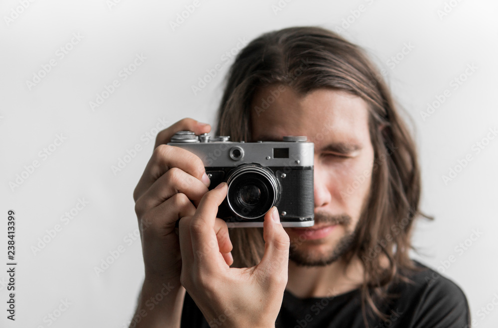 Handsome young bearded man with a long hair and in a black shirt holding vintage old-fashioned film camera on a white background and looking in camera viewfinder. Handsome young bearded man with a long hair and in a black shirt holding vintage old-fashioned film camera on a white background and looking in camera viewfinder.