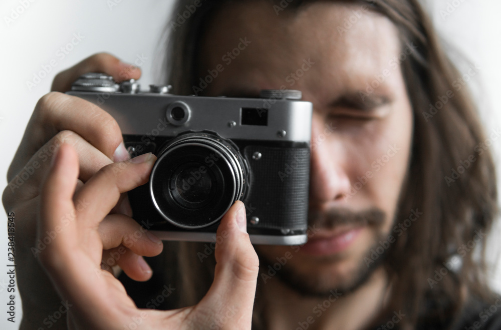 Handsome young bearded man with a long hair and in a black shirt holding vintage old-fashioned film camera on a white background and looking in camera viewfinder. Handsome young bearded man with a long hair and in a black shirt holding vintage old-fashioned film camera on a white background and looking in camera viewfinder.