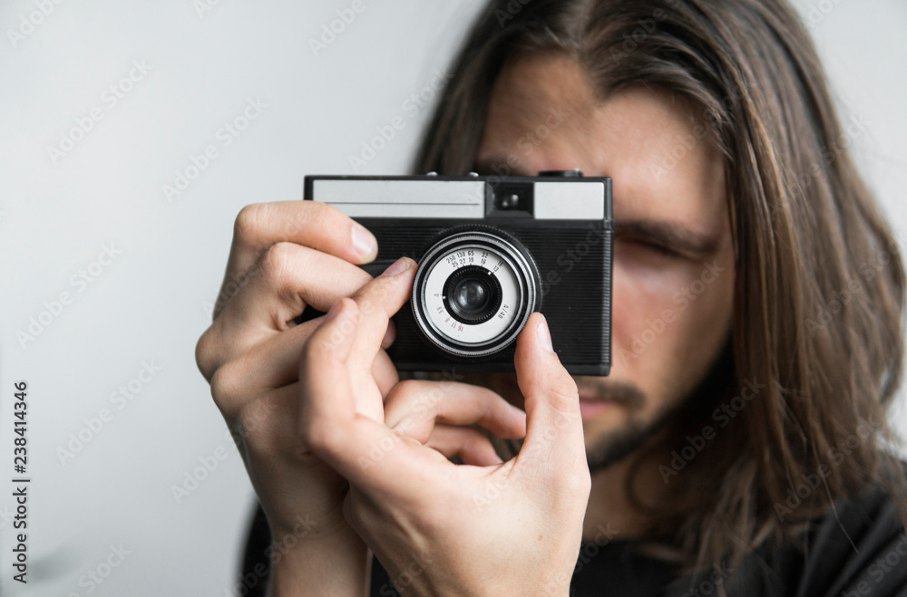 Handsome young bearded man with a long hair and in a black shirt holding vintage old-fashioned film camera on a white background and looking in camera viewfinder. Handsome young bearded man with a long hair and in a black shirt holding vintage old-fashioned film camera on a white background and looking in camera viewfinder.