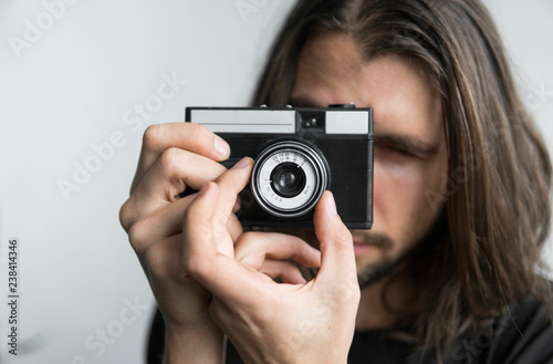 Handsome young bearded man with a long hair and in a black shirt holding vintage old-fashioned film camera on a white background and looking in camera viewfinder. © Volodymyr