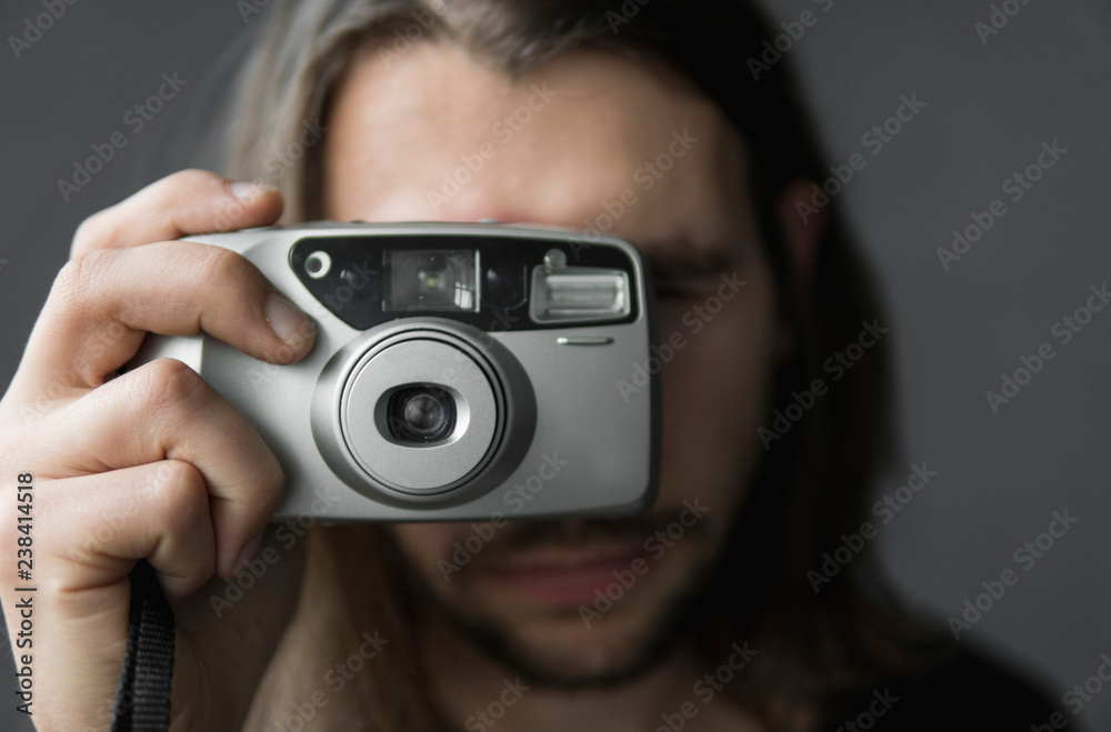 Handsome young bearded man with a long hair and in a black shirt holding vintage old-fashioned film camera on a black background and looking in camera viewfinder. Handsome young bearded man with a long hair and in a black shirt holding vintage old-fashioned film camera on a black background and looking in camera viewfinder.