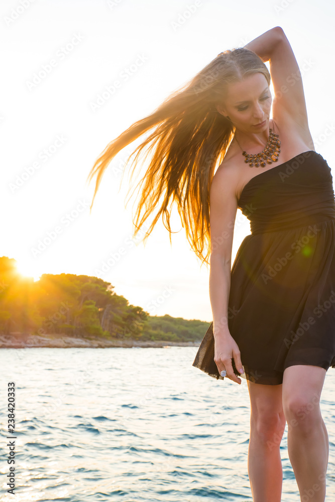 Naklejka premium A beautiful woman in an elegant black dress standing and posing on the rocks next to the sea during the sunset.