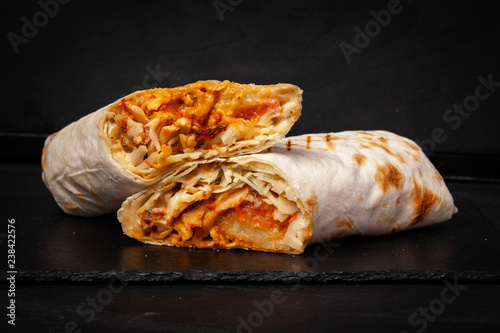 Shawarma sandwich gyro- fresh roll of thin lavash (pita bread) filled with grilled meat, mushrooms, cheese, cabbage, carrots, sauce, green. Traditional Eastern snack. On dark black background