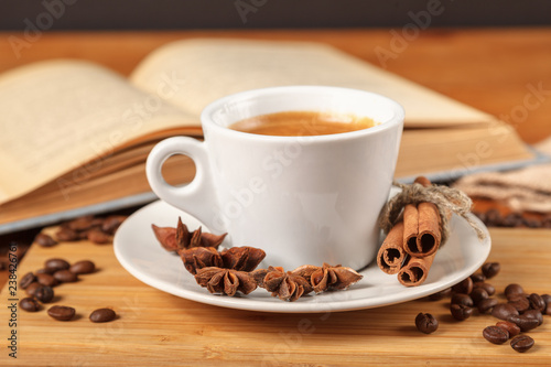 Coffee break a Cup of hot coffee and a book on a wooden table. A white Cup of black coffee surrounded by a small amount of roasted coffee beans, cinnamon, anise and an open book