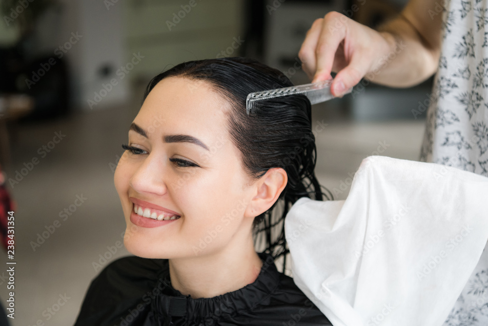 Fototapeta premium Close up of a young happy beautiful woman smiling to the camera while professional hairdresser wrapping her wet hair in a towel after washing it with shampoo