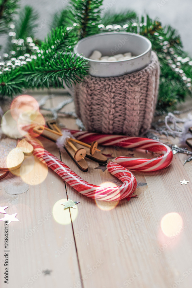 White cup for cocoa in beige knitted clothes. mittens on a white wooden background with christmas tree and red caramel candy White cup for cocoa in beige knitted clothes. mittens on a white wooden background with christmas tree and red caramel candy