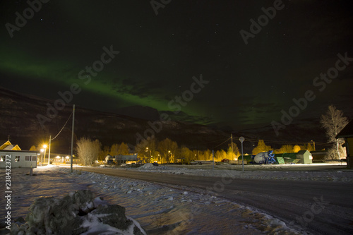 Fototapeta Naklejka Na Ścianę i Meble -  The northern lights (aurora borealis) rising behind a mountain in a small village in the north of Norway. In the middle of winter with snow covering the streets.
