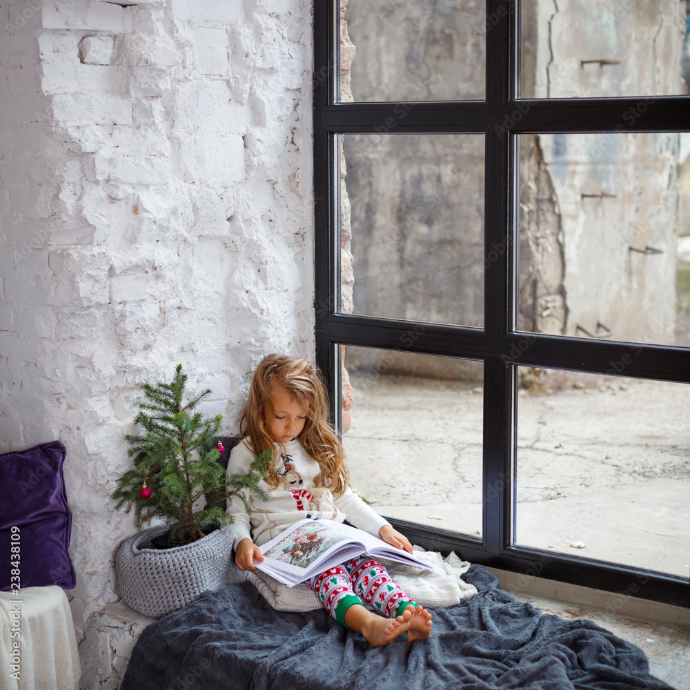 Portrait of little pretty girl reading book and sitting on windowsill near Christmas tree in cozy room. Cute girl with her gift on Christmas