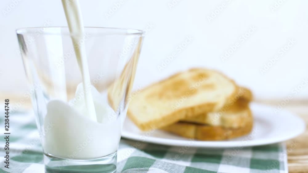 Milk pouring in glass with toast bread on white plate and green tablecloth on wooden kitchen surface. Slow motion footage. Milk pouring in glass with toast bread on white plate and green tablecloth on wooden kitchen surface. Slow motion footage.