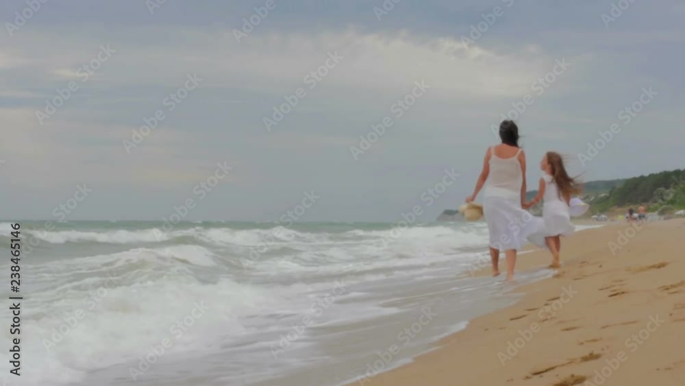mother walks with her daughter along the beach holding hands, in white dresses with hats in her hands mother walks with her daughter along the beach holding hands, in white dresses with hats in her hands