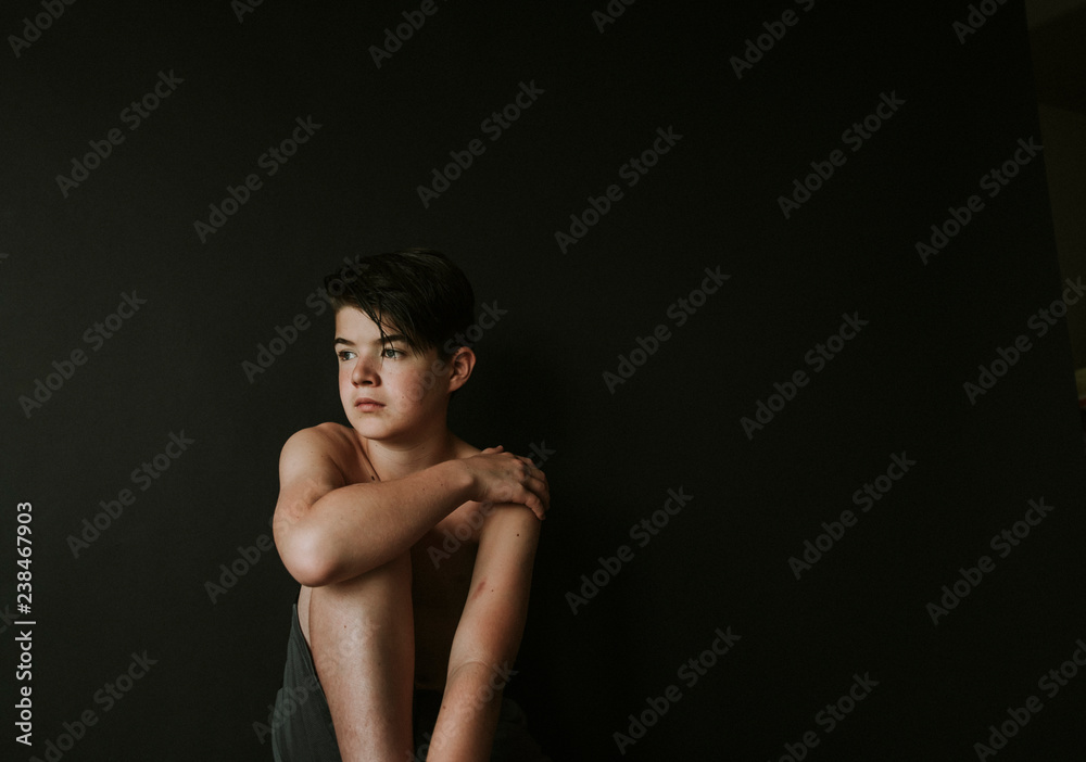 Thoughtful shirtless boy looking away while sitting against black background Thoughtful shirtless boy looking away while sitting against black background