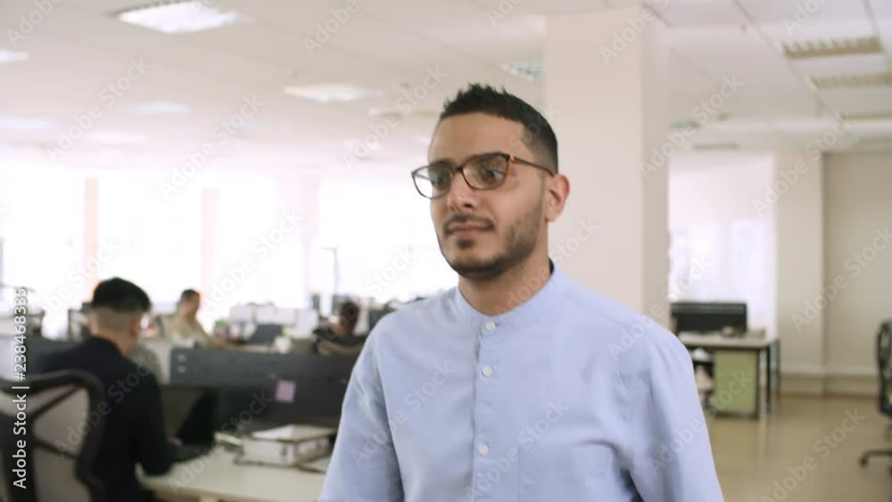Front view medium shot of young Asian man in glasses walking through modern office and smiling Front view medium shot of young Asian man in glasses walking through modern office and smiling