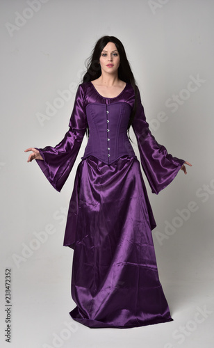 full length portrait of beautiful girl with long black hair,   wearing purple fantasy medieval gown. standing pose on grey studio background.