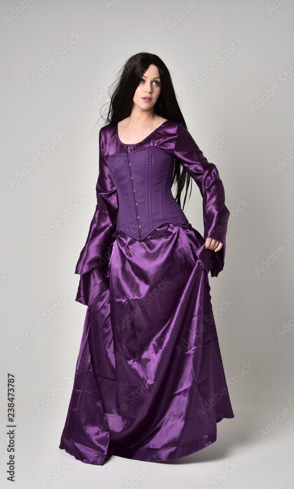 full length portrait of beautiful girl with long black hair, wearing purple fantasy medieval gown. standing pose on grey studio background. full length portrait of beautiful girl with long black hair, wearing purple fantasy medieval gown. standing pose on grey studio background.