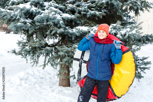 cheerful cute young boy in hat red scarf and blue jacket holds tube on snow, has fun, smiles. Teenager on sledding in winter park. Active lifestyle, winter activity, outdoor winter games © Natali