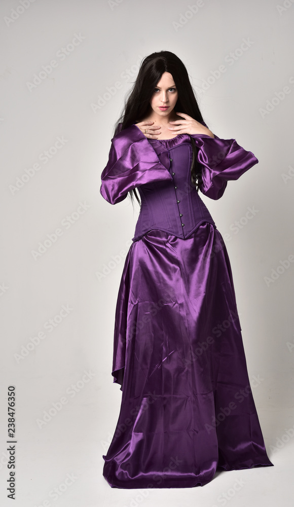 full length portrait of beautiful girl with long black hair, wearing purple fantasy medieval gown. standing pose on grey studio background. full length portrait of beautiful girl with long black hair, wearing purple fantasy medieval gown. standing pose on grey studio background.