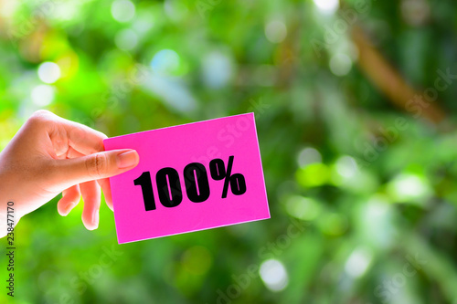 A woman's hand holding a pink color paper with a text '100%' written on it. Green nature background. © SURIYATI