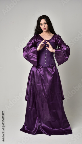 full length portrait of beautiful girl with long black hair,   wearing purple fantasy medieval gown. standing pose on grey studio background.