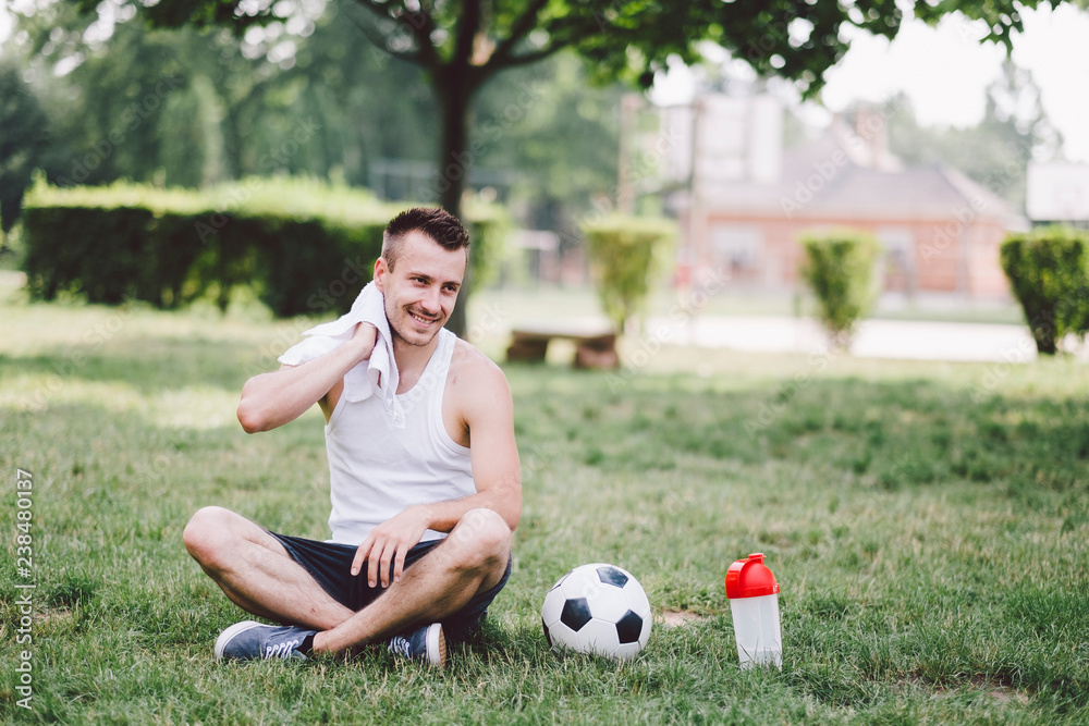 Smiling young man wiping neck while sitting on soccer field at park Smiling young man wiping neck while sitting on soccer field at park