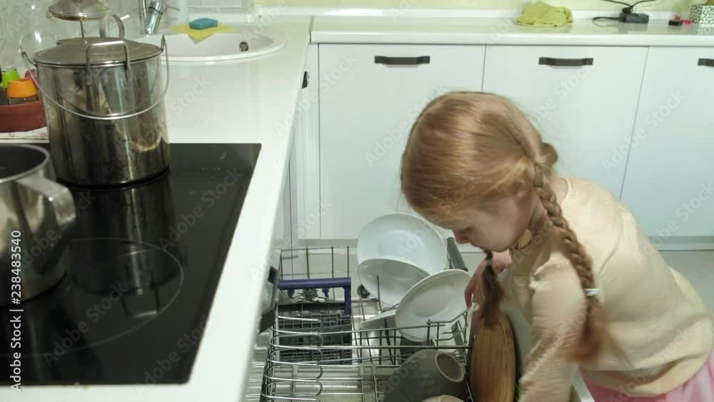 Cute blonde babe girl in a pink skirt, helps mom in the bright kitchen, loads plates, cups in the dishwasher Cute blonde babe girl in a pink skirt, helps mom in the bright kitchen, loads plates, cups in the dishwasher