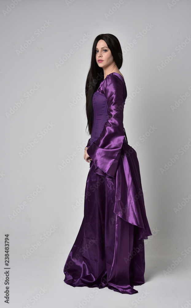 full length portrait of beautiful girl with long black hair, wearing purple fantasy medieval gown. standing pose on grey studio background. full length portrait of beautiful girl with long black hair, wearing purple fantasy medieval gown. standing pose on grey studio background.