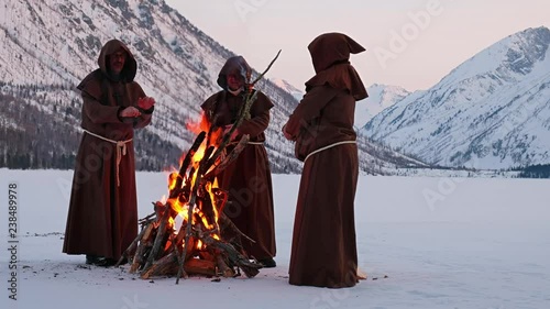 Group of monks in brown cloaks of the order of St. Francis are warming themselves by the fire