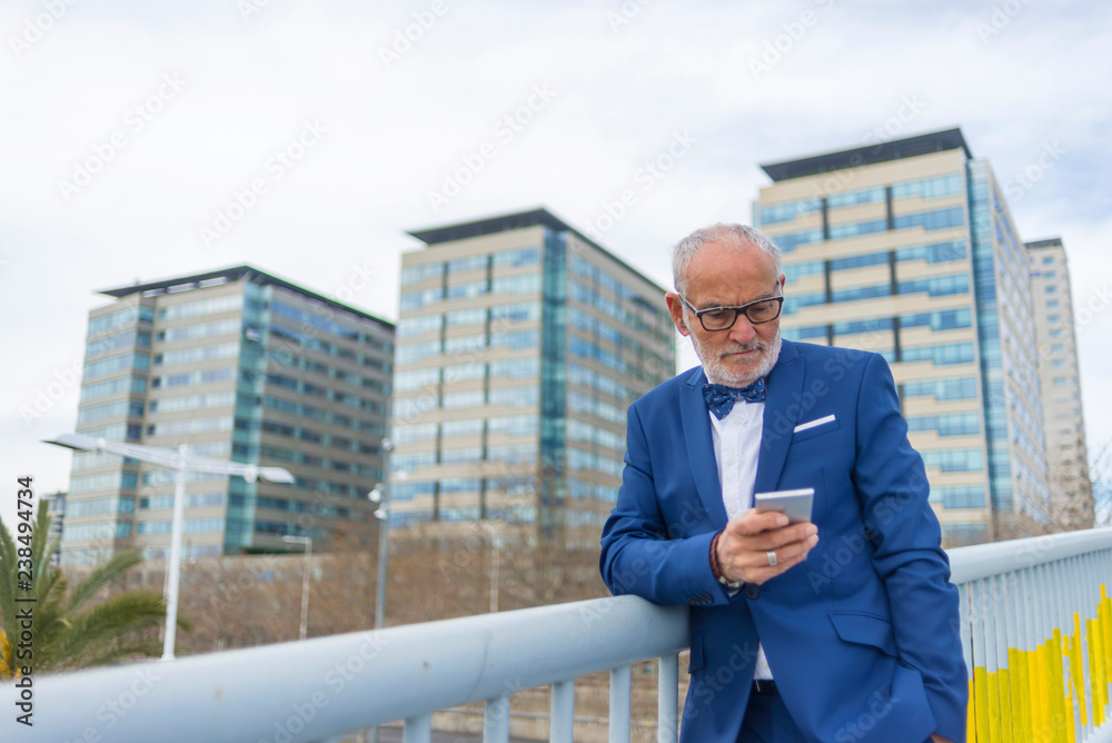 Low angle view of confident senior man using smart phone while standing by railing against sky in city Low angle view of confident senior man using smart phone while standing by railing against sky in city