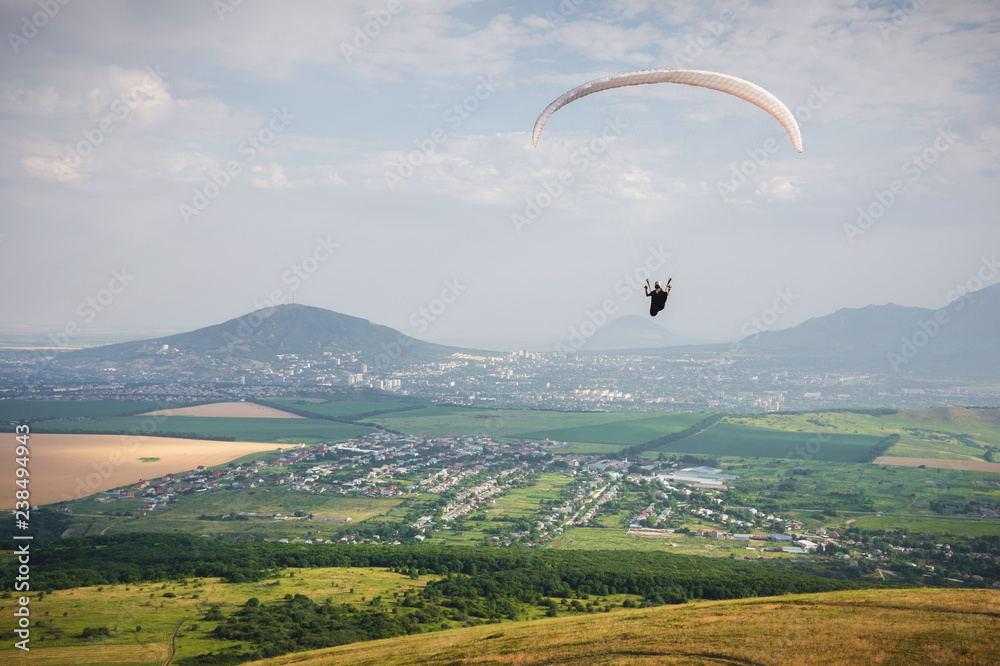 Professional paraglider in a cocoon suit flies high above the ground against the sky and fields with mountains Professional paraglider in a cocoon suit flies high above the ground against the sky and fields with mountains