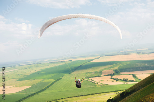 Professional paraglider in a cocoon suit flies high above the ground against the sky and fields