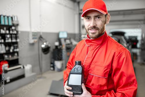 Portrait of a handsome auto mechanic in red uniform holding bottle with engine oil at the car service