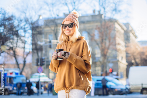 Blond positive hipster stylish finn woman in beige hat and hoody, warm mittens drinks coffee from disposable cup in winter time outdoor.