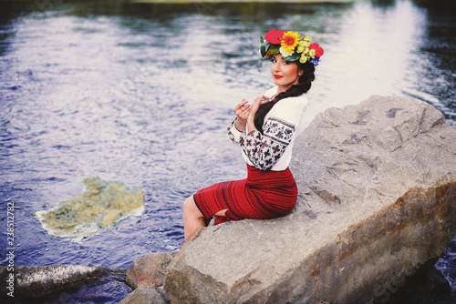 Fototapeta Naklejka Na Ścianę i Meble -  Beautiful woman with dark long hair in national dress red skirt and a white shirt sitting on the stone near river