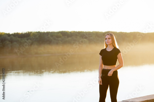 Fitness woman wearing black top and leggings and stands on the background of rocks on the river embankment