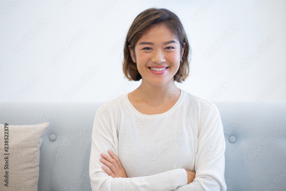 Portrait of smiling young woman looking at camera. Cheerful Asian girl sitting on coach with folded arms. Beauty concept Portrait of smiling young woman looking at camera. Cheerful Asian girl sitting on coach with folded arms. Beauty concept