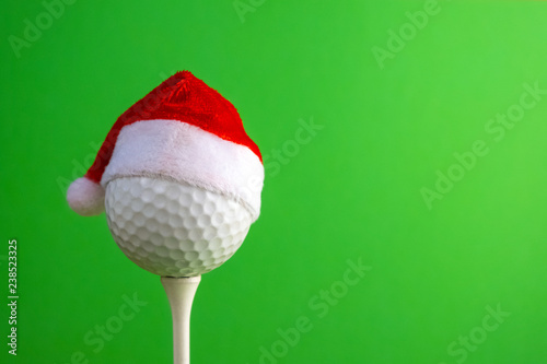 Sports concept on the topic of golf, Christmas and New Year. White golf-ball in a red Santa Claus hat set on a tee. Green background. Free space.