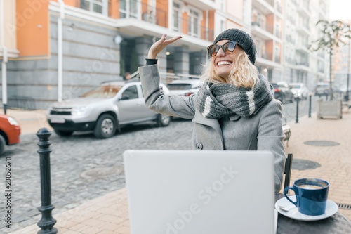 Young woman blogger freelancer in warm clothes hat in outdoor cafe with computer laptop, mobile phone, city street background