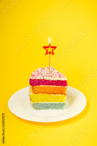 Slice of Rainbow cake with birning candle in the shape of star on white round plate isolated on bright yellow background. Happy bithday, party concept. Vertical card. Selective focus. Copy space.