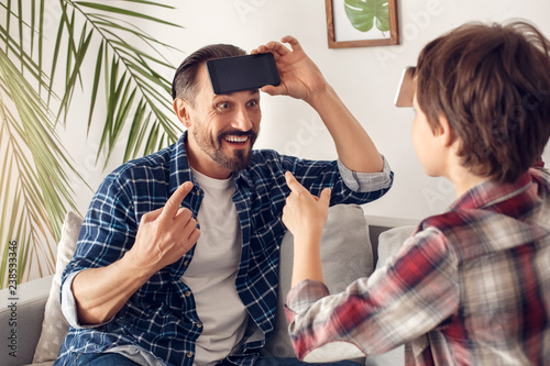 Father and little son at home sitting on sofa playing heads up with smartphone guessing word cheerful photo