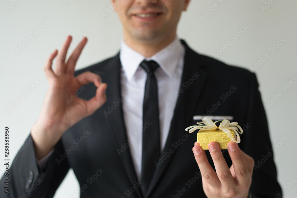 Close-up of happy businessman holding small gift box and showing ok sign. Smiling man in suit advising to buy Christmas gift. Choice concept Close-up of happy businessman holding small gift box and showing ok sign. Smiling man in suit advising to buy Christmas gift. Choice concept