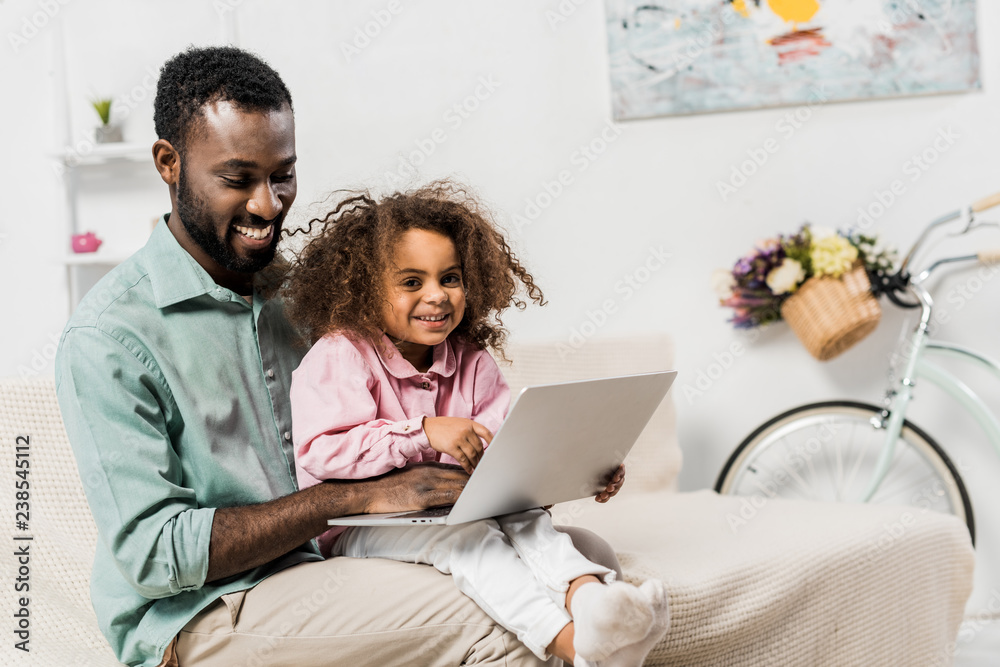 african american father and adorable child sitting on couch and using laptop african american father and adorable child sitting on couch and using laptop