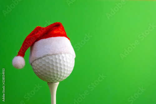 Golf ball on a tee with a red santa claus hat. Template for design greeting card for golfer for new year or christmas. Green background. Copyspace.