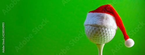 The golf ball mounted on the tee is wearing a Santa Claus souvenir hat. Blank for a postcard for a golfer to celebrate the New Year and Christmas. Bright green background. Copy space.
